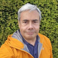 A grey haired man standing in front of a green bush wearing a yellow jacket.