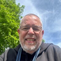 A smiling man with a beard and glasses wearing a grey top