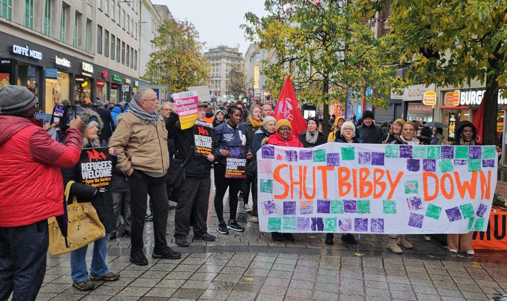 A group of people standing in a high street holding a banner saying Shut Bibby Down