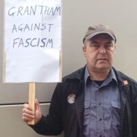 A man wearing a peak cap, blue shirt and black jacket holding a sign saying Grantham against Fascism.